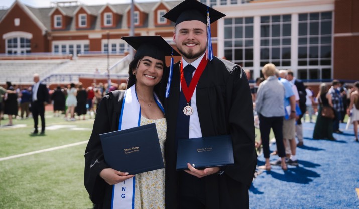Female and male student smiling and posing with their diplomas on football field